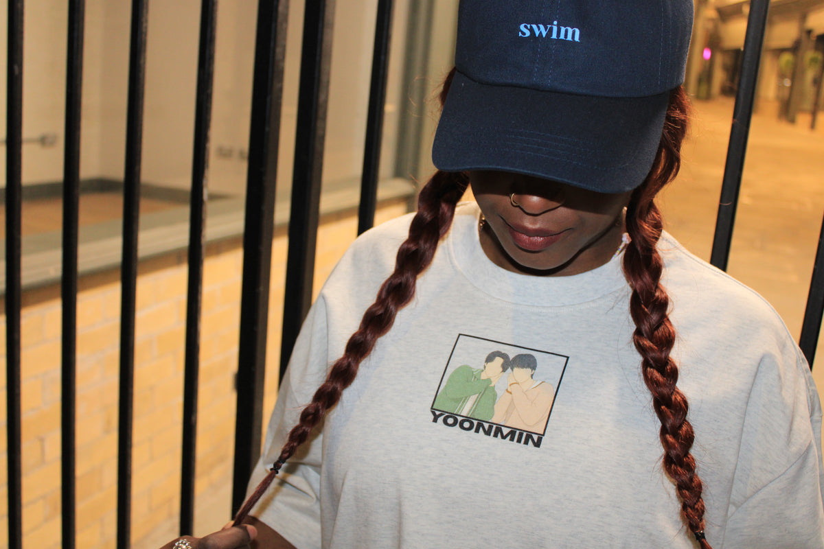 A person wearing a navy blue baseball cap with the word 'swim' and a light gray sweatshirt featuring a graphic print with the name 'YOONMIN'.