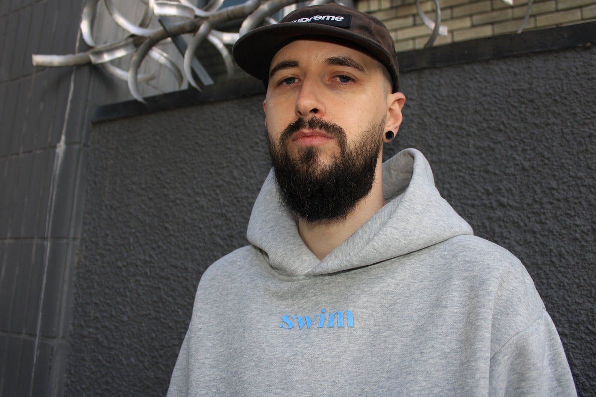 A man with a beard and a black Supreme cap wearing a light gray hoodie with the word 'swift' printed in light blue on the chest, standing against a dark gray textured wall.